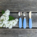 Stenblå børnebestik i silikone og rustfrit stål ved siden af hvide blomster på træbord.