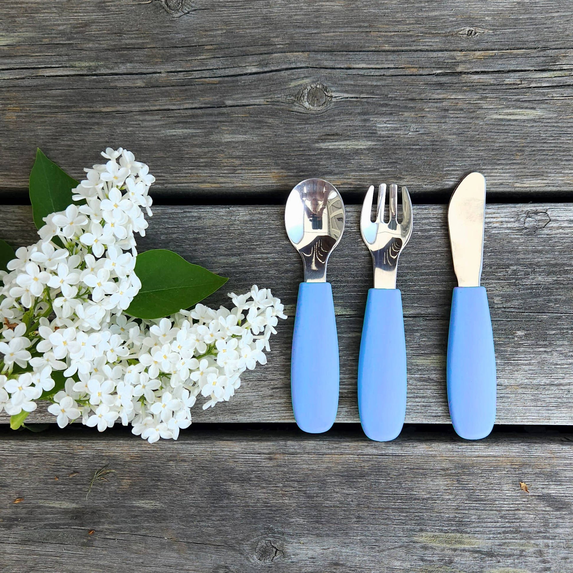 Stenblå børnebestik i silikone og rustfrit stål ved siden af hvide blomster på træbord.