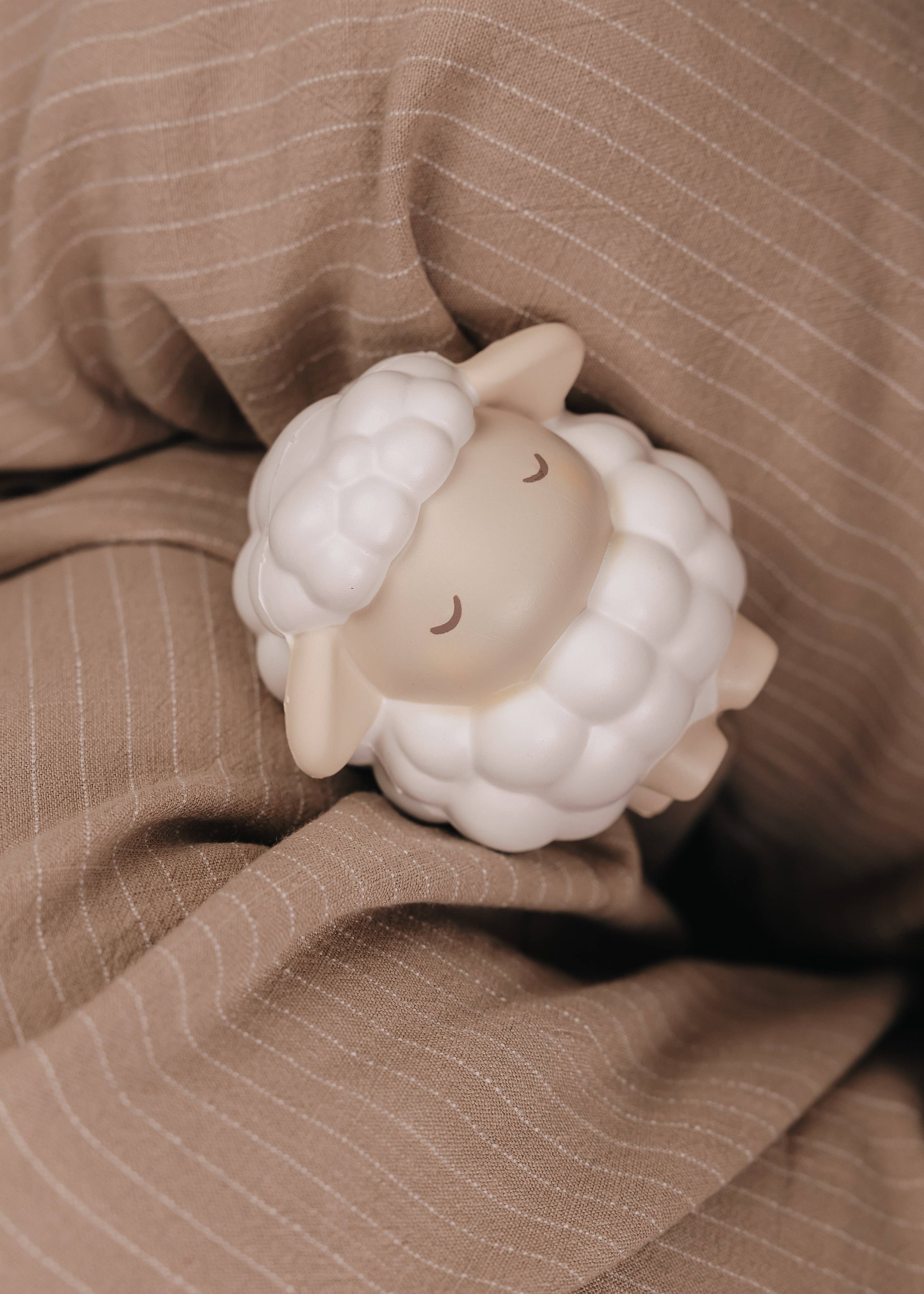 Et hvidt og beige Squichymal uldne fåret legetøj i blødt skum liggende på brun tekstil.