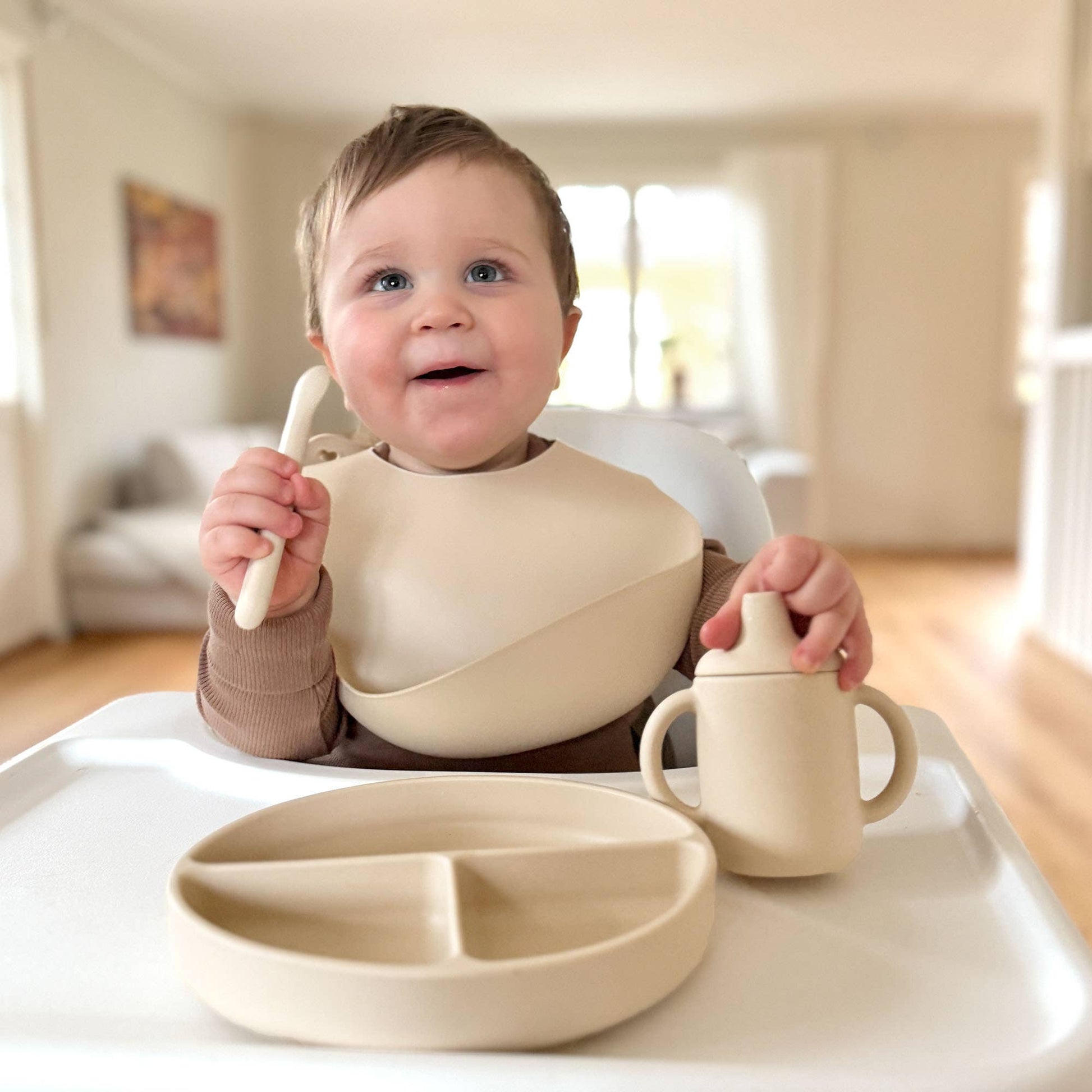 Glad baby ved spisebord med opdelt silikone tallerken, hagesmæk og tudkop i farven Sandshell.
