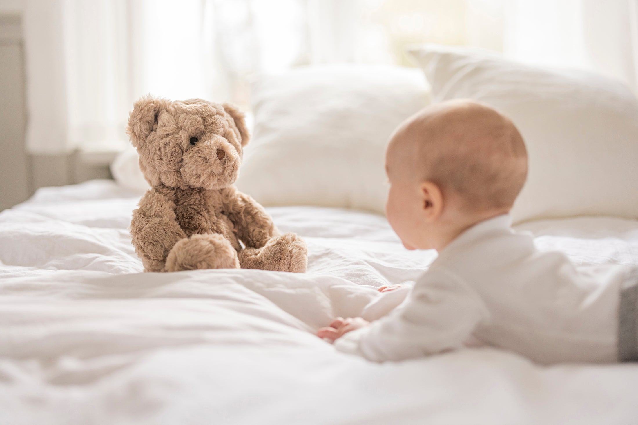 En baby der ligger på en hvid seng og kigger på bamsen Sture.