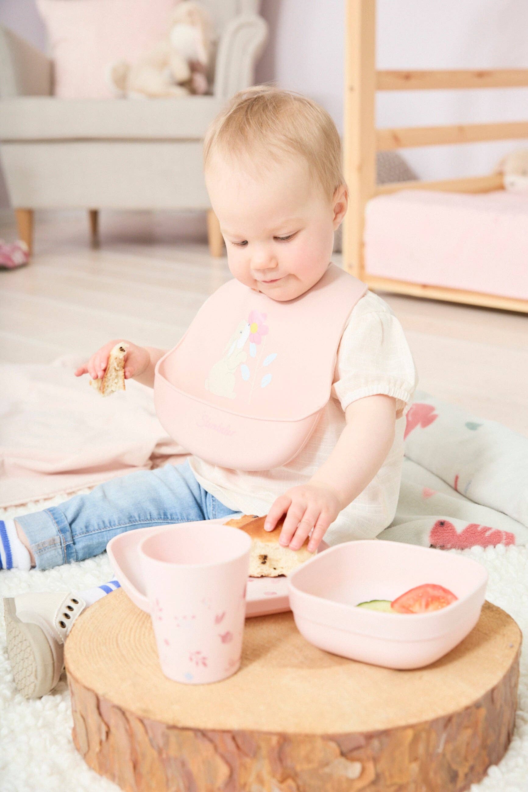 Baby sidder med lyserød Kanin Hanni silikonehagesmæk og spiser ved et lille bord.