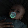 Baby med BIBS GLOW sut i mørket, grøn ring lyser, forældrehånd holder baby