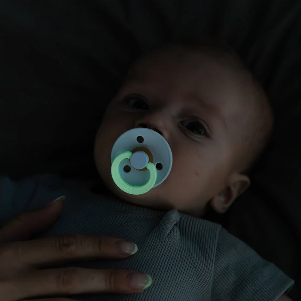 Baby med BIBS GLOW sut i mørket, grøn ring lyser, forældrehånd holder baby