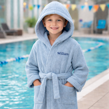 Barn i lyseblå badekåbe med broderet navn ved en swimmingpool.