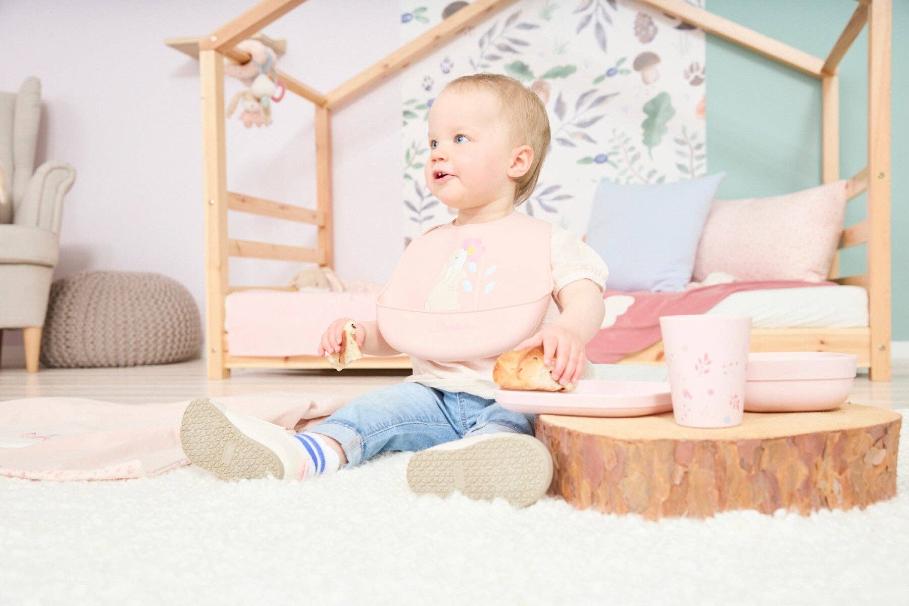 Baby med lyserød hagesmæk sidder på gulvet og spiser med Sterntaler Hase Hanni servicesæt.