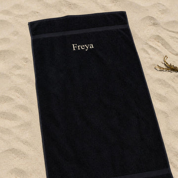 Sort personligt håndklæde med navnet Freya broderet, liggende på en sandstrand.