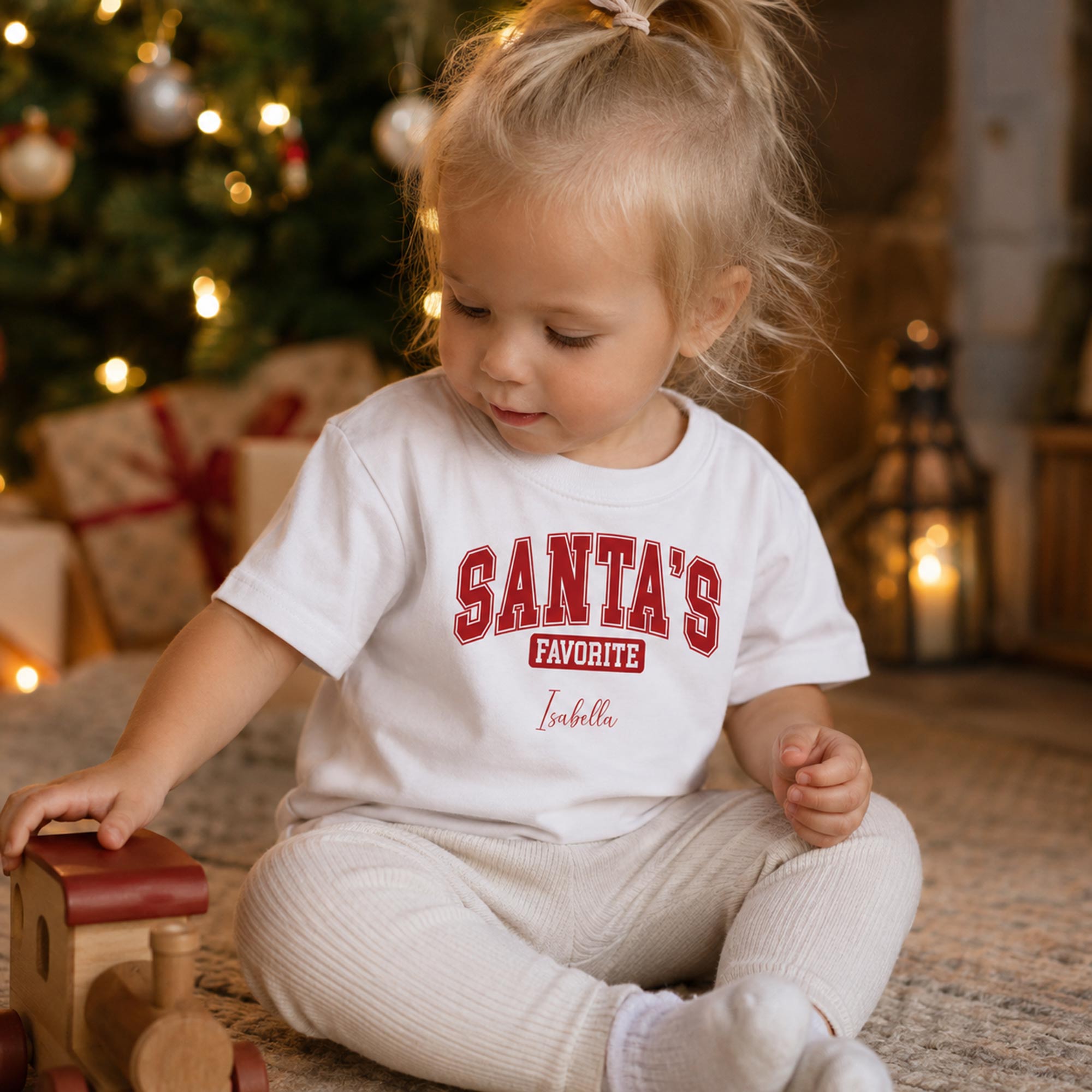 Lille pige i hvid Santas Favorite t-shirt leger foran et juletræ med gaver.