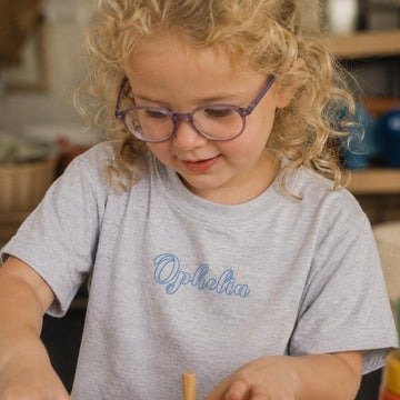 Lille pige med briller i lysegrå t-shirt med navnet Ophelia trykt på brystet.